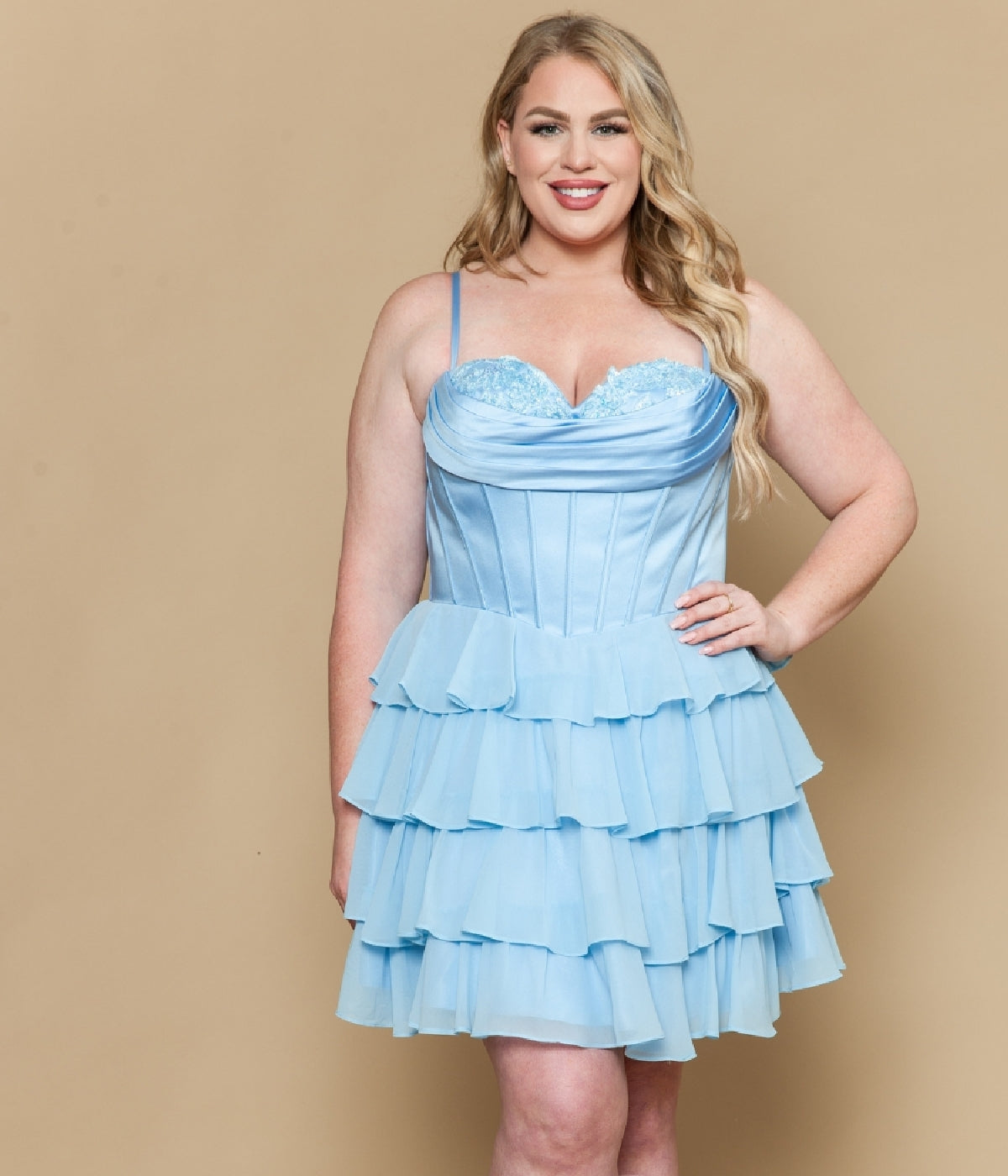 A woman wearing the Short Plus Size Homecoming Dress: Poly USA W1196—a light blue, spaghetti strap dress with a satin corset bodice and tiered ruffle details—smiles at the camera against a beige background.